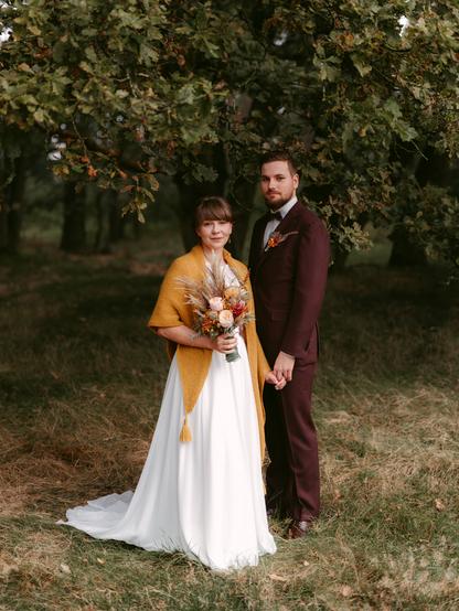 This is a beautiful autumn wedding portrait featuring a couple standing together in a natural outdoor setting. The bride is wearing a flowing white wedding dress with a distinctive mustard yellow knitted shawl or wrap draped over her shoulders. She has a short bob haircut with bangs and is holding a rustic autumn bouquet featuring dried grasses, orange and burgundy flowers, and natural textures.  The groom stands beside her wearing a rich burgundy or maroon suit with a black bow tie and a matching floral boutonniere on his lapel. He has brown hair and a well-groomed beard.  They're positioned on a patch of dried grass beneath the canopy of a large tree with autumn foliage - the leaves showing beautiful fall colors of green, yellow, and orange. The background is softly blurred, creating a dreamy, intimate atmosphere typical of professional wedding photography.  The lighting appears to be natural, golden hour light that gives the entire scene a warm, romantic glow. The couple is holding hands and both are looking directly at the camera with gentle, content expressions. The overall aesthetic is very much in line with modern, intimate elopement photography - natural, authentic, and beautifully composed.