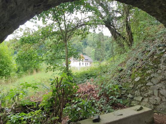 Der Blick fällt unter einem Torbogen hindurch. Der steinerne Bogen ist Teil der Drachenbahnstrecke. Man schaut hindurch auf ein weißes Haus, welches etwas weiter weg einzeln auf einer Wiese steht und von Bäumen umgeben ist.