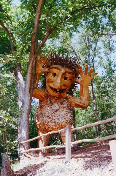 A series of large wooden troll and animal sculptures made from recycled materials, located in a forest setting in Detroit Lakes, Minnesota. The expressive, oversized figures- some sitting, some standing, and one bright yellow rabbit - are part of an outdoor art installation by Danish artist Thomas Dambo, blending whimsy with environmental commentary. Lush greenery surrounds the sculptures, creating a surreal, fairy-tale-like atmosphere.
