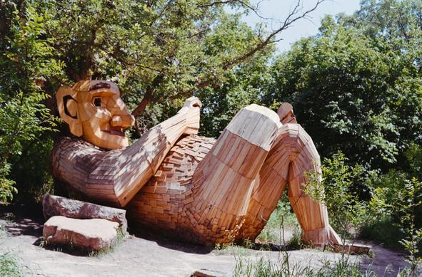 A series of large wooden troll and animal sculptures made from recycled materials, located in a forest setting in Detroit Lakes, Minnesota. The expressive, oversized figures- some sitting, some standing, and one bright yellow rabbit - are part of an outdoor art installation by Danish artist Thomas Dambo, blending whimsy with environmental commentary. Lush greenery surrounds the sculptures, creating a surreal, fairy-tale-like atmosphere.
