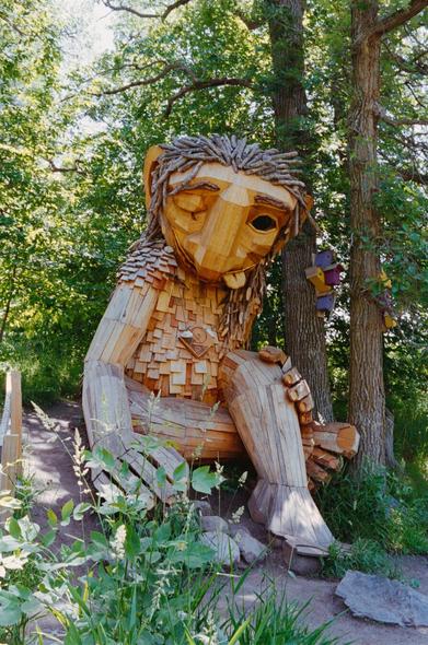 A series of large wooden troll and animal sculptures made from recycled materials, located in a forest setting in Detroit Lakes, Minnesota. The expressive, oversized figures- some sitting, some standing, and one bright yellow rabbit - are part of an outdoor art installation by Danish artist Thomas Dambo, blending whimsy with environmental commentary. Lush greenery surrounds the sculptures, creating a surreal, fairy-tale-like atmosphere.