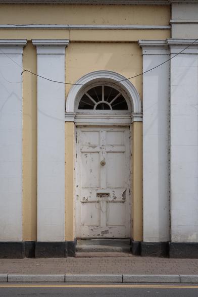 Cenrally framed in a Georgian-style facade with flat columns - an old, solid, windowless door with peeling white paint, a single round brass door handle at centre two thirds of the way up, a letterbox that's inexplicably spewing white cabling - beneath a small arched window. At the side of a street, with a brick-tiled path passing right in front of the door, concrete kerbstones and a double yellow line on the tarmac.