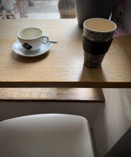 Eine weiße Tasse mit dem Logo des Cafés & ein eigener Kaffeebecher stehen auf einer breiten Fensterbank, davor ist unscharf die Sitzfläche eines weißen Stuhls zu sehen