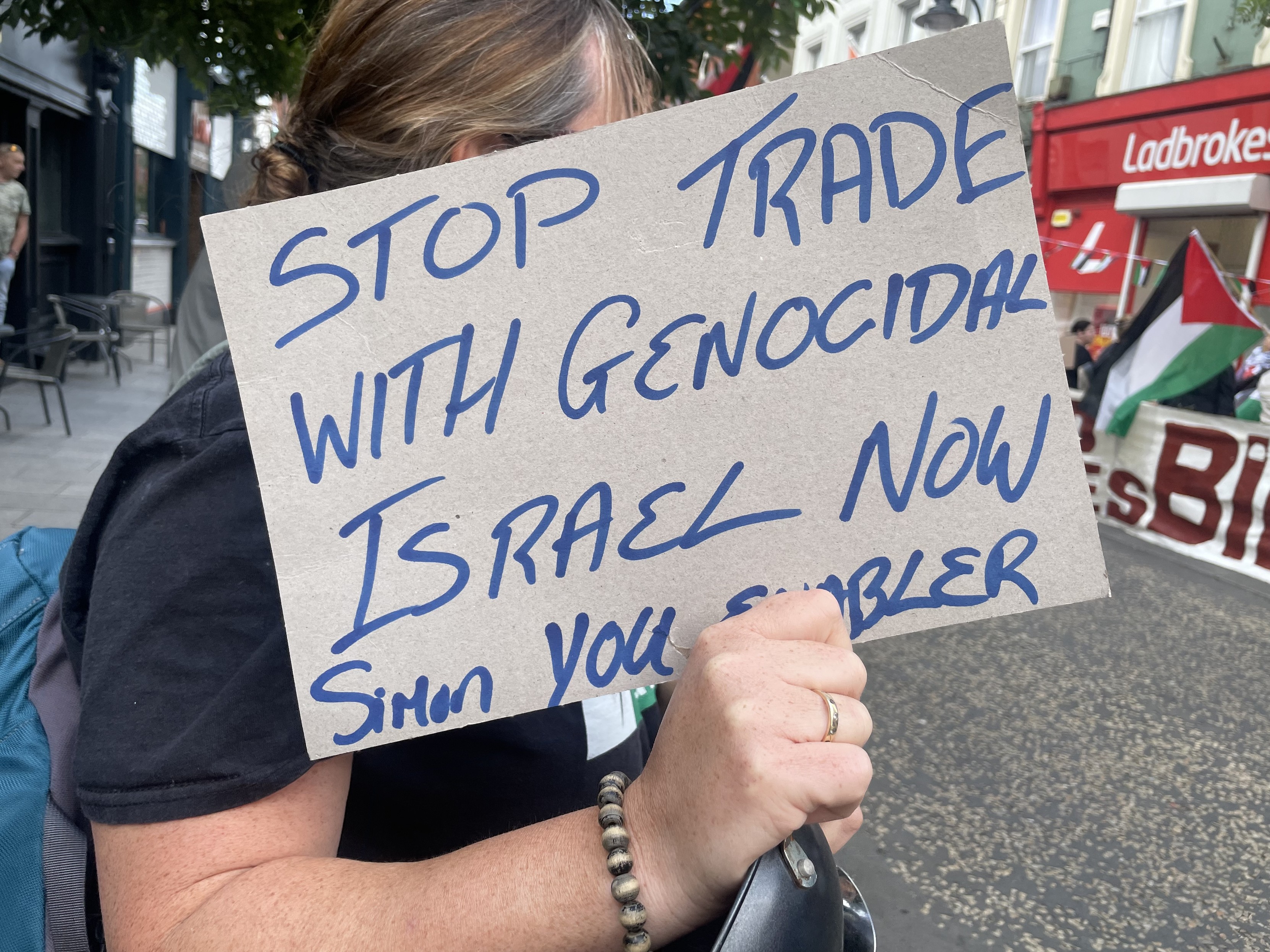 Protestor with sign “stop trade with genocidal Israel now, Simon you enabler”