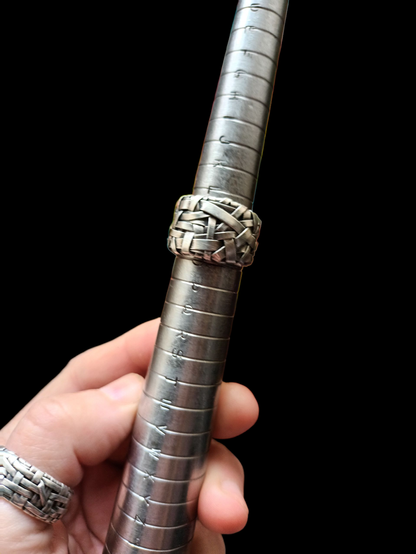 A hand holding a ring sizer with a woven silver ring on it,in a vlack backfeound. Another woven silver ring band on the thumb.