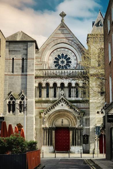A frontal, eye-level photo of St Ann's Church in Dublin. The large, grey stone church facade dominates the centre of the image, featuring a deep red wooden main door under a Romanesque arch. Above the door are several arched windows, and a large, ornate circular stained-glass window sits at the very top. The church is flanked on both sides by taller brick buildings, and the ground is a paved street with a small plant pot and orange parasols visible on the left. The sky is partially cloudy with patches of blue visible.