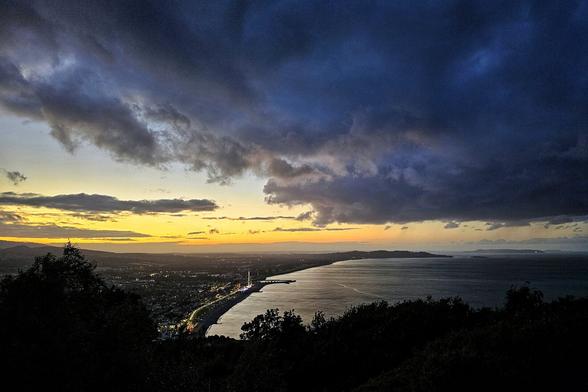 A dramatic, wide-angle landscape photo taken from a high vantage point overlooking a coastal city at dusk. The sky is dominated by dark, heavy storm clouds on the left, transitioning to a bright, golden-orange glow on the horizon to the right. Below, the sea reflects the dim light, and the city lights of Bray, County Wicklow, twinkle along the curving coastline. The foreground is silhouetted with dark trees, adding to the moody atmosphere.