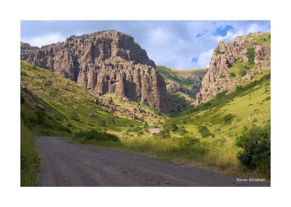 The image depicts a winding road that leads to jagged mountains on either side of it. There are green hills with little tree coverage in the foreground, and background.