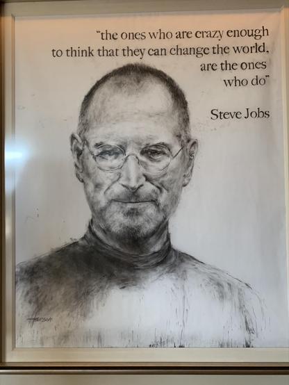 Black and white charcoal sketch of a middle-aged man wearing glasses and a turtleneck, framed and displayed on a white wall. Above the portrait is a quote attributed to Steve Jobs: “the ones who are crazy enough to think that they can change the world, are the ones who do.” The artist's signature is in the bottom left corner of the sketch.