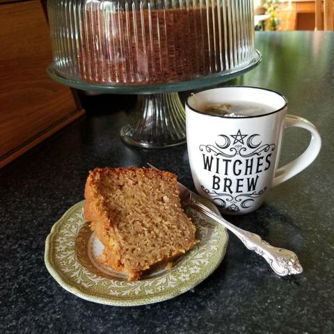 On a kitchen counter sits a small plate with an unadorned slice of pumpkin spice cake, a cup of tea with "witches brew" on the mug, and a glass plate stand with the rest of the cake inside the glass cover.