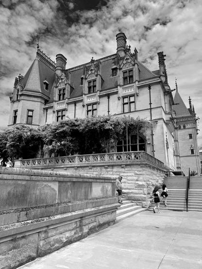 A black and white image of the Biltmore House which is very ornate and resembles European palaces and castles. Located in Asheville, North Carolina in the US, it is owned by the Vanderbilt family, although they no longer reside in the house, but nearby. Public tours help pay for the upkeep. People are walking up stairs to the front of the house.