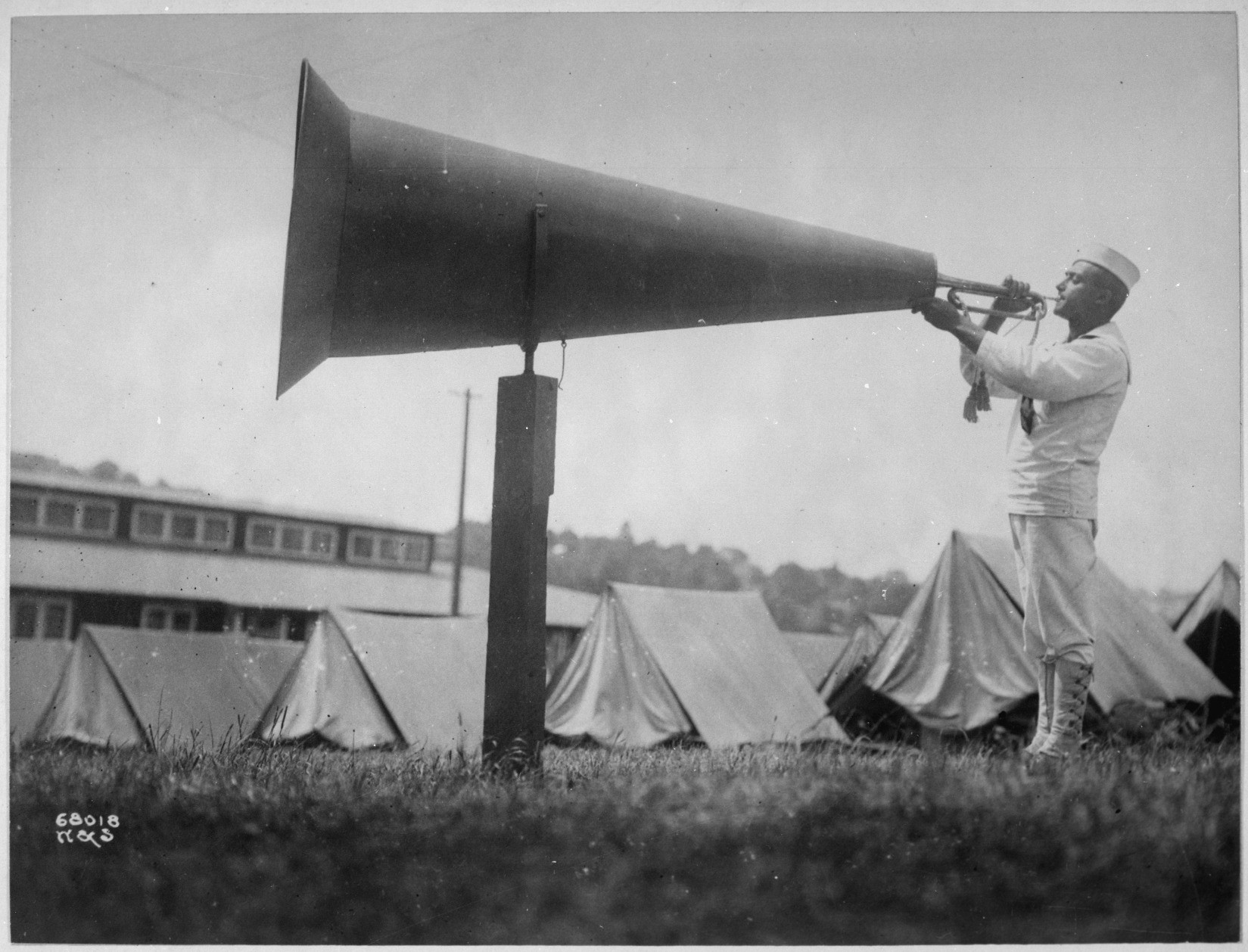 black and white photo of a man in navy whites playing a bugle into a colossal megaphone – seriously, this megaphone is 7 or 8 feet long with an opening at the far end of about four feet (it of course is mounted on a stand). in the background there are tents which presumably house the men being awakened momentarily by reveille