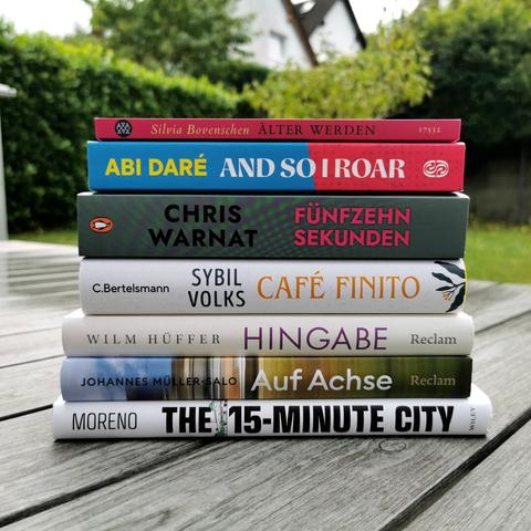 Auf dem Foto ist ein aus sieben Büchern bestehender Bücherstapel auf einem gräulichen Holztisch. Im Hintergrund sind ein Garten mit Hecke und weiße Häuser zu erkennen. Die Bücher werden in der Caption aufgelistet.