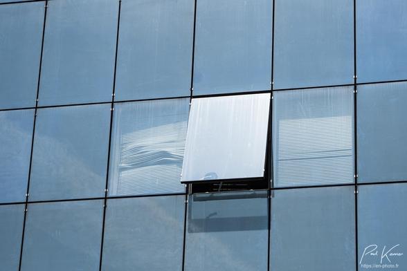 Photographie en couleur présentant le reflet du ciel, très peu nuageux, sur la façade intégralement en verre d'un immeuble de bureau. La couleur dominante est bien sûr le bleu sur les plaques de verre séparées par des joints en caoutchouc noir. Le ciel se reflète sur la quasi totalité du verre à l'exception de trois plaques, pratiquement au centre de la photo : à travers deux d'entre elles on voit le store vénitien abaissé et plus ou moins abîmé alors qu'entre ces deux plaques, une plaque-fenêtre basculante est entrouverte vers le bas, avec par conséquent,  un reflet plus clair du fait de l'angle différent. Le bas et la poignée de cette fenêtre se reflètent sous la plaque de verre située juste en dessous.