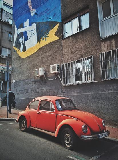 A vertical, full-color, eye-level shot captures a bright red vintage Volkswagen Beetle parked on a city street. The car occupies the bottom half of the frame, angled slightly toward the viewer. In the background, a large building wall is visible, featuring a mural of a figure with yellow hair and blue cloth, alongside text. Below the mural, a window with metal bars and two air conditioning units are mounted on the wall. A man in a dark jacket walks away from the camera on the left side of the street. The car's license plate, partially visible, reads "BG 578-ŠJ." The lighting is bright and even.