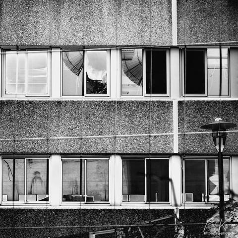 Photographie monochrome en noir et blanc de format carré, présentant deux étages de fenêtres d'un immeuble de bureau en cours de démolition, certaines fenêtres sont brisées, alors qu'à travers d'autres fenêtres, on aperçoit des stores vénitiens mal-en-point, en éventail…