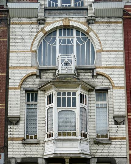 Partie d’une façade Art nouveau en brique blanche avec une logette en bois blanc qui a l’air un peu vieillotte, flanquée de deux étroites fenêtres. Une jolie baie vitrée arrondie se trouve juste au-dessus.