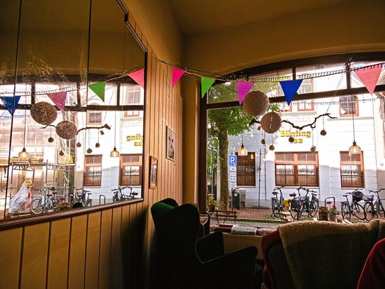 This photo was taken in one of my favourite cafés in Leer, called ‘Rusty Rooster’. My gaze falls out of the window onto a bright house across the street, on which the words ‘Bünting Tee’ are written in golden letters. The sun shines on it. Various bicycles are parked in front of the house. The interior of the café is painted light brown, and on the left wall is a mirror reflecting the house opposite. In front of the window, inside, is a green armchair, and the back of a red sofa with a blanket over it can be seen. It is a peaceful place, very cosy.