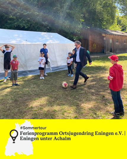Minister Manne Lucha spielt Fußball mit Kindern auf einer Wiese vor einem Zelt und einem Holzhaus bei sonnigem Wetter. Text: Ferienprogramm Ortsjugendring Eningen e.V., Eningen unter Achalm.
