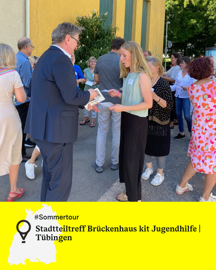 Minister Manne Lucha übergibt einer jungen Frau ein Dokument bei einer Veranstaltung im Freien, sie sind umgeben von weiteren Personen. Text: Stadtteiltreff Brückenhaus kit Jugendhilfe, Tübingen.