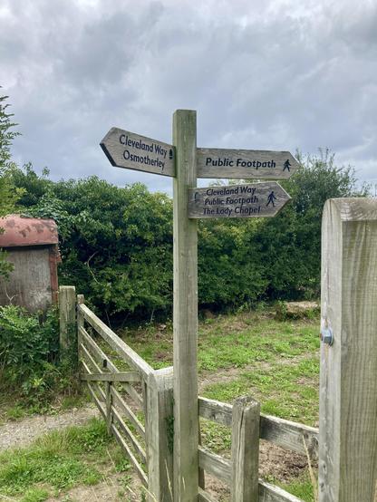 Photo of a wooden fingerpost with three arms. Two are showing the route along the Cleveland Way and the other marks a public footpath. The fingerpost is right next to a wooden fence and gate that marked the boundary between a farm and fields. In the background are small tress and bushes and an old corrugated shed. It's a cloudy, overcast day.