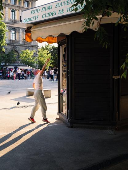 A young woman with pink hair pulls down the rolling shutter of her souvenir kiosk at sunset. The low sun projects long shadows.