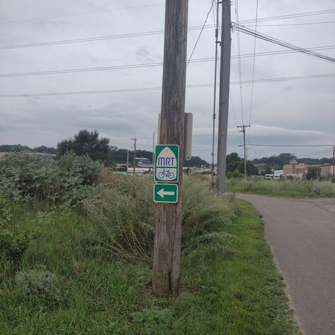 Green and White sign for the Mississippi River Trail, or MRT, up on a wooden utility pole. A greenway path goes through an industrial area the background.