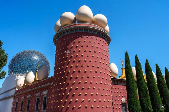 Color photo of a round tower at the corner of a building with giant white eggs on the top. The left side of the building has a large glass geodesic sphere on top and to the right is a row of skinny cypress trees in front of the building.  The walls of the building are deep burgundy-red color and covered with golden ornaments that look like bread rolls.
