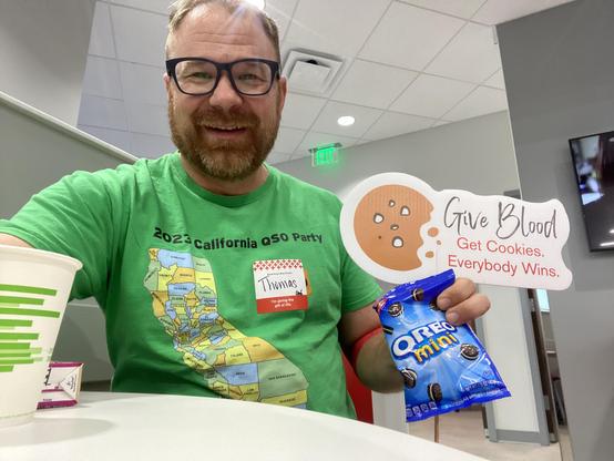 A man in a green shirt holds a bag of mini Oreos and a sign that says Give Blood get cookies everybody wi s