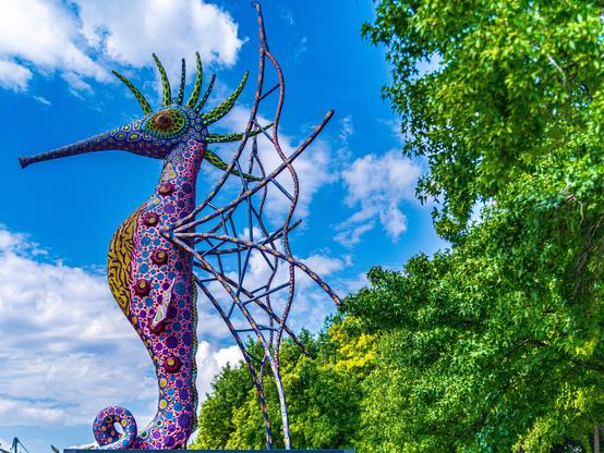 A colorful sculpture of a seahorse, featuring intricate patterns in shades of purple, green, and orange, is set against a bright blue sky with clouds and lush green trees in the background.

Kolorowa rzeźba konika morskiego, z misternymi wzorami w odcieniach fioletu, zieleni i pomarańczy, jest osadzona na tle jasnoniebieskiego nieba z chmurami i bujnymi zielonymi drzewami w tle.