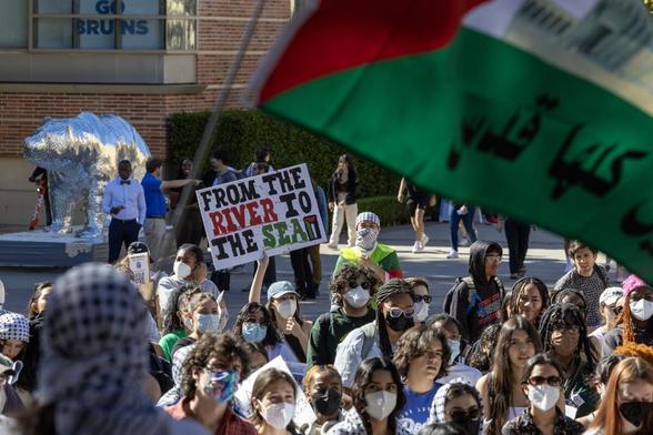 Campus protest sign seen saying "From The River To The Sea" amidst crowd and a Palestinian Flag seen in support of Palestinians outside a UCLA bldg