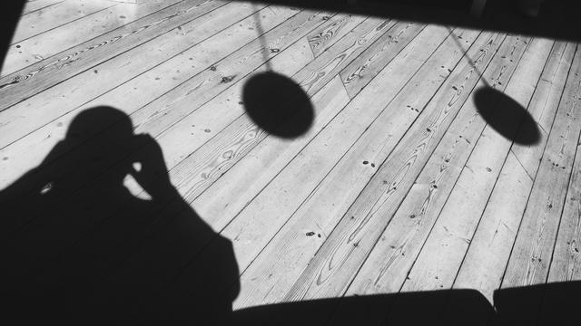 Wooden floor in bright sunlight, with stark shadows from two globe shaped lights hanging, and the photographer, cast sharply in the floor