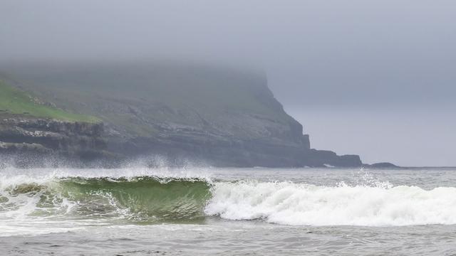A foggy coastal scene featuring green hills and rocky cliffs on the left. Waves crash gently on the shore, with a misty atmosphere creating a tranquil mood.