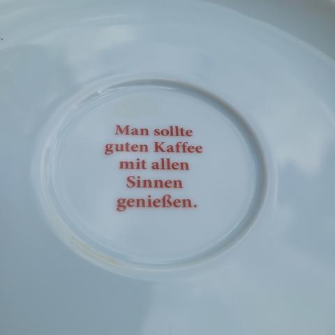 Weißer Kaffeeunterteller. In der Mitte steht:man sollte guten Kaffee mit allen Sinnen genießen