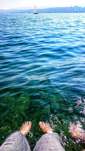 A first-person point of view shot looking out over a large body of water. The viewer's legs, wearing light grey denim trousers, are visible at the bottom of the frame. Their bare feet are submerged in the clear, rippling water, which transitions from a deep blue in the distance to a translucent, turquoise green near the shore, revealing the rocky lakebed below. The surface of the water sparkles in the sunlight. In the distance, several sailing boats are scattered across the water, and a hazy shoreline with hills is visible on the horizon.