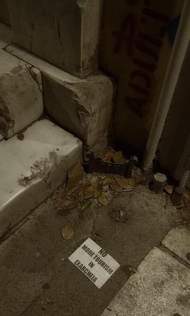Detail of dirty city pavement.
Diagonal areas of shades and lights. 
Top left, partially seen, the broken marble steps of an entrance. 
Beside them, surrounded by fallen leaves, a horizontal piece of ironwork fixed in concrete. It's a podómactron. 
It indicates that this is quite an old house, since its purpose was to remove mud from shoes, when the streets were still unpaved. 
The word ADULT is stenciled vertically on the wall above.
A white flyer with bold black capital letters demands: no more tourism in Exarcheia.