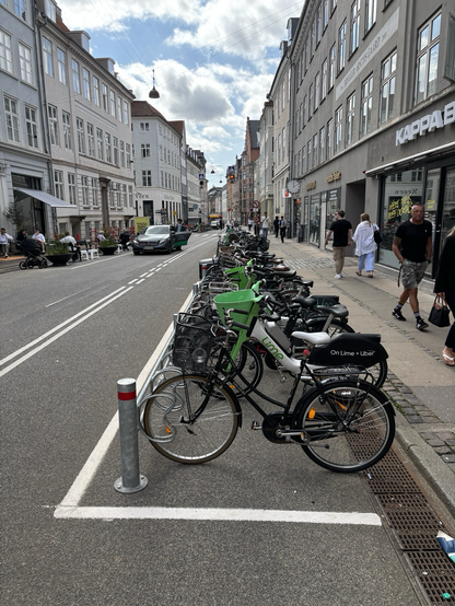 Eine fast autoleere Straße mit breiten Radstreifen. Rechts eine große Anzahl Fahrräder die statt Autoparkplatz dort abgestellt sind. Links sitzen Menschen auf der Straße vor einem Cafe, an einer Stelle, wo vorher Autoparkplätze waren.
