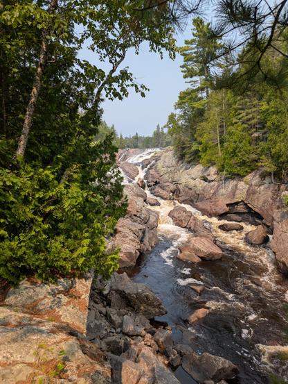 A photo of a roaring waterfall and river through a channel of igneous rock. Blue sky and sunshine. Cedar trees line both sides of the river.