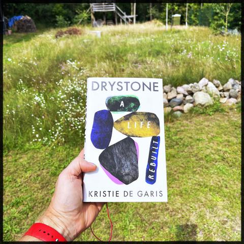 A left hand holding a book in front of a garden. The garden is mostly meadow and grass and small cobble stone wall. The book is "Drystone - A life Rebuilt" from Kristie De Garis.