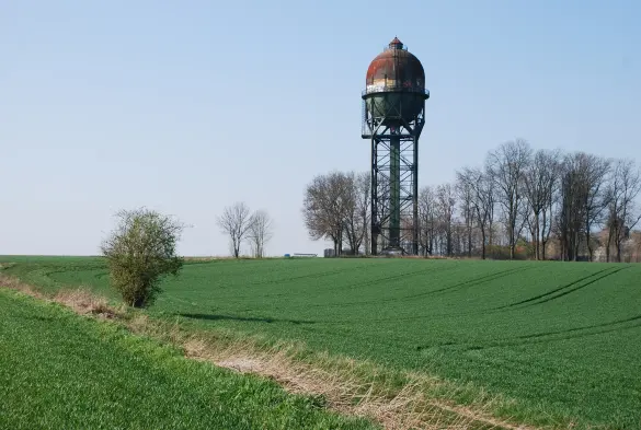 The image shows a water tower standing tall in a green field. The water tower has a rounded top with a rusty appearance, and it is supported by a tall metal structure. In the foreground, there is a path leading through the field, and a few trees are visible nearby. The sky is clear and blue, indicating a sunny day. The overall scene conveys a rural landscape.

Image Credits: Wikimedia / Rkikultur / CC BY-SA 4.0