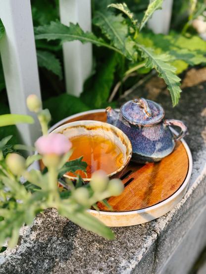 A blue and purple glazed teapot and teacup on a bamboo tray sitting on a concrete wall. Along the wall is a white fence where weeds have taken over. A purple thistle flower is in bloom but blurred in the foreground.