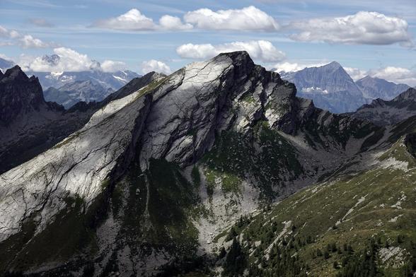 Im Vordergrund ist ein felsiger, schroffer und teils grasiger Berggipfel zu sehen, im Hintergrund ist eine weitere Bergkette mit Schneefeldern zu sehen, die teils in Wolken gehüllt sind.