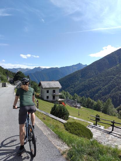 Im Vordergrund ist ein Radfahrer auf einem Gravelbike zu sehen, der auf einer ins Tal abfallende Straße unterwegs ist. Rechts sind die Ausläufer eines kleinen Alpendorfes mit steileren Wiesen und Wald zu sehen. Im Hintergrund sind größtenteils bewaldete Bergketten erkennbar.