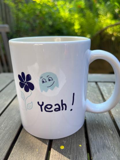 Weiße Henkeltasse. Auf der Tasse steht Yeah, daneben eine Blume und ein Geist.