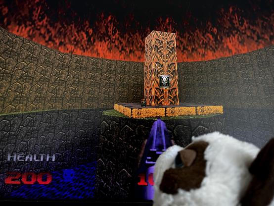English: Another moment from the game Doom 64. Again it shows a round recess in the level, the floor of which is covered with water textures, with a dark brown wall rising in a semicircle at its edges, the lower half of which is finished off with a narrow, dark gray textured ridge. In the center is a platform with a pillar on it, which is designed in a lighter brown color with decorations reminiscent of H.R. Giger, and a switch is embedded in it. 
The flames of hellfire can be seen as the sky again.
In the foreground is the head of a stuffed cat, now photographed in such a way that it looks as if the cat was looking suspicious or angry at the game.

Deutsch: Ein weiterer Moment aus dem Spiel Doom 64. Erneut ist eine runde Ausbuchtung im Level zu sehen, deren Boden von Wassertexturen bedeckt ist, an dessen Rändern sich im Halbkreis eine dunkelbraune Wand erhebt, die in der unteren Hälfte noch von einem schmalen und dunkelgrau texturierten Grat abgeschlossen wird. In der Mitte ist eine Plattform, auf der ein Pfeiler steht, der in hellerem braun und mit an H.R. Giger erinnernde Verzierungen gestaltet ist, ist ein Schalter eingelassen.
Als Himmel sind auch hier wieder die Flammen des Höllenfeuers zu sehen.
Im Vordergrund ist der Kopf der Plüschkatze nun aber so zu sehen, dass es aussieht, als würde die Katze das Spiel misstrauisch oder mürrisch beobachten.