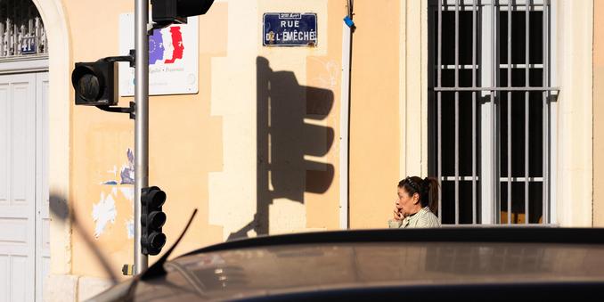 Street scene with a woman on a phone near a building marked 'Rue Delémeche' in France. A traffic light casts a shadow on the beige wall. A car roof is partially visible in the foreground. A window with metal bars is slightly open on the right. Signs and posters are affixed to the wall.