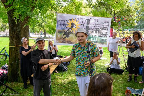 Die Kriegsdienstverweigerer Konstantin aus der Ukraine und Andrej aus Russland geben sich die Hand. Bei einem Konzert des Protestorchesters Lebenslaute am Denkmal des Deserteurs in Potsdam. Am 80ten Jahrestag des Atombombenabwurfs auf Nagasaki. Potsdam, 09.08.2025