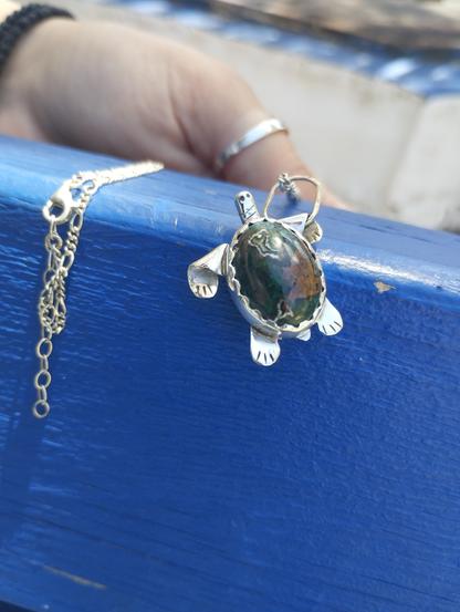 a pendant in shape of a turtle. it's silver with oval green gemstone. It's placed on blue background