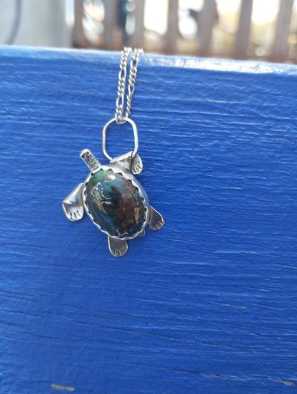 a pendant in shape of a turtle. it's silver with oval green gemstone. It's placed on blue background