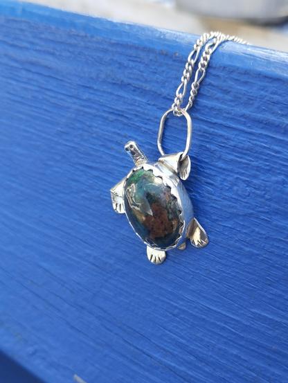 another view of the pendant in shape of a turtle. it's silver with oval green gemstone. It's placed on blue background